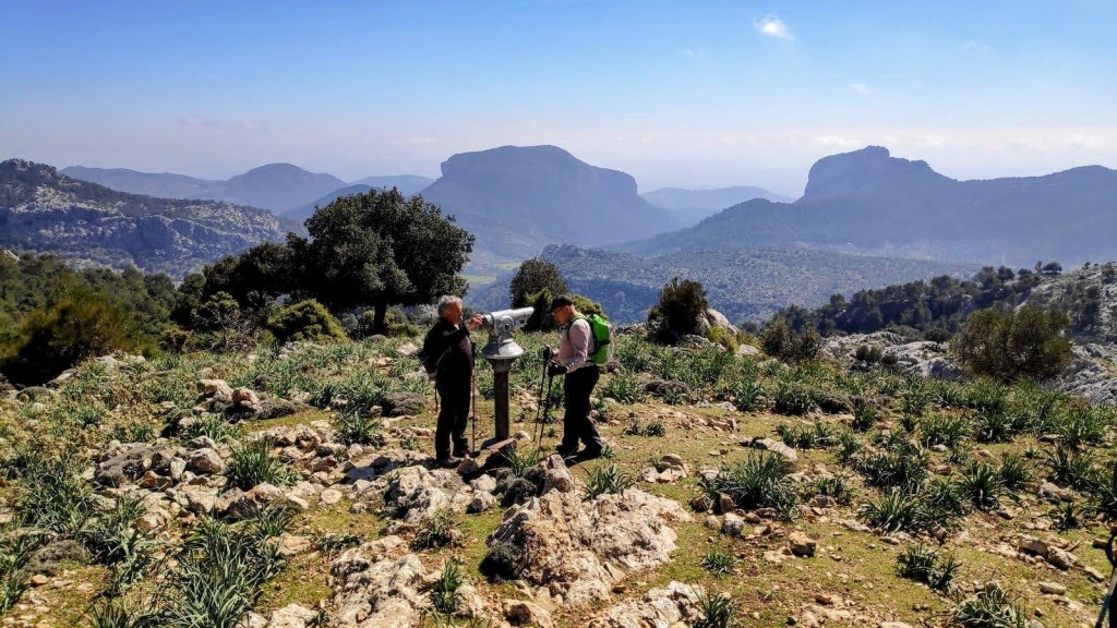 Dos excursionistas observando el paisaje desde un mirador del Coll d'en Poma, con montañas y un valle visible al fondo.