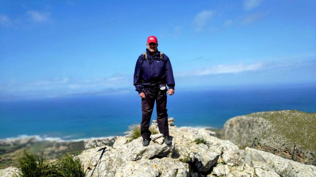 Hombre de pie sobre rocas en la cima del Bec de Ferrutx, con vistas al mar y paisaje montañoso de fondo.