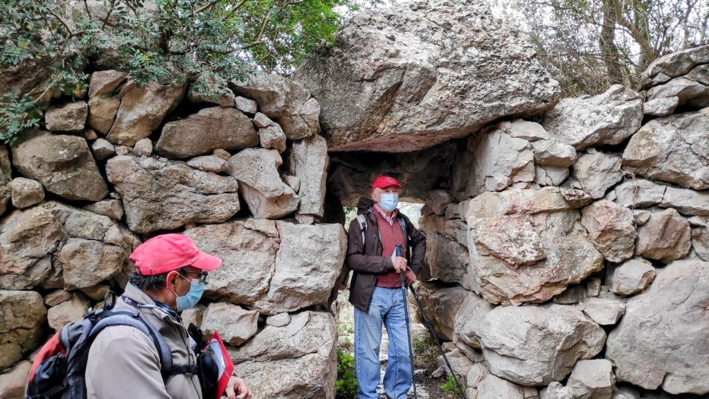 Dos hombres con gorros rojos y mascarillas de protección, de pie frente a una entrada estrecha de Talaiot de Son Coll Nou, en un entorno natural.