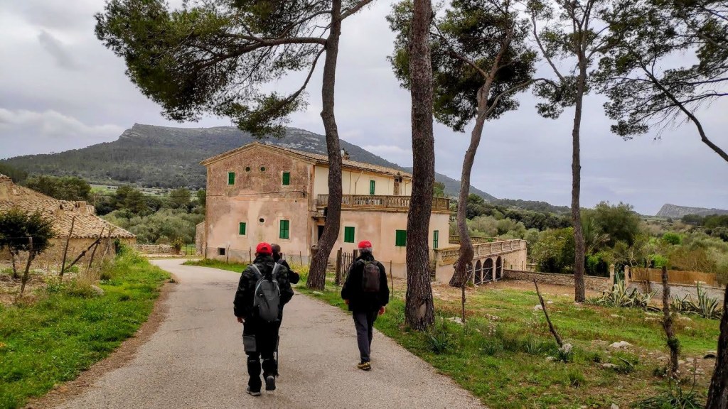 Dos senderistas caminando por un camino rural, rodeados de árboles y vistas a montañas, acercándose a las Casas de Castellitx, una casa de estilo rural con ventanas verdes.