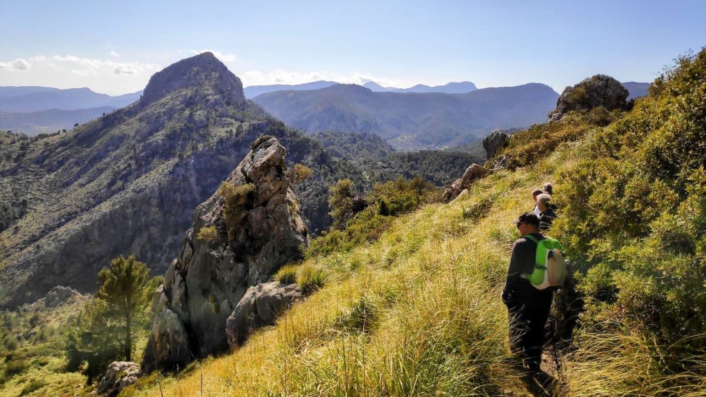 Senderistas caminando por un sendero montañoso con vegetación verde y rocosas; se observa un paisaje montañoso de fondo.