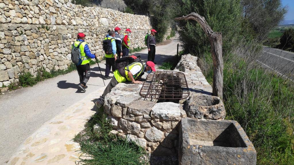 Grupo de excursionistas observando un pozo tradicional en un camino rural en Mallorca, rodeados de vegetación y una pared de piedra.