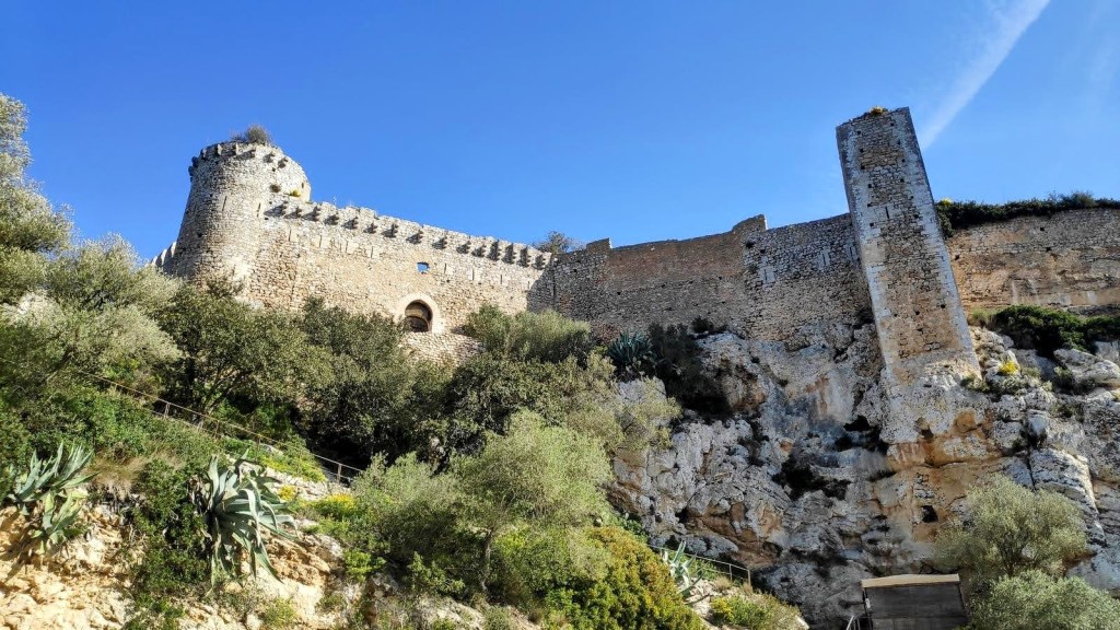 Vista del Castillo de Santueri, un castillo en ruinas situado sobre un acantilado, rodeado de vegetación y con un cielo azul despejado al fondo.