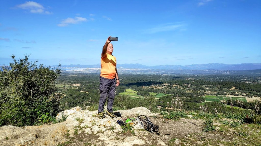 Una persona de pie en una roca, tomando una selfie con un teléfono móvil, mientras se aprecia un paisaje verde y montañoso al fondo bajo un cielo despejado.