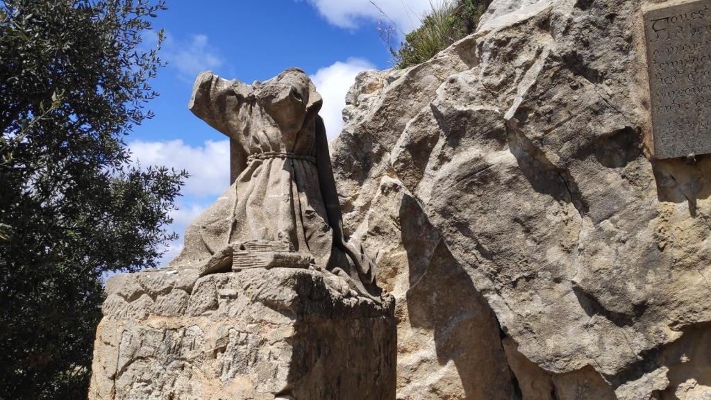 Estatua desgastada  del Beato Ramón Llullen, un pedestal de piedra, rodeada de rocas y vegetación, bajo un cielo azul con nubes.