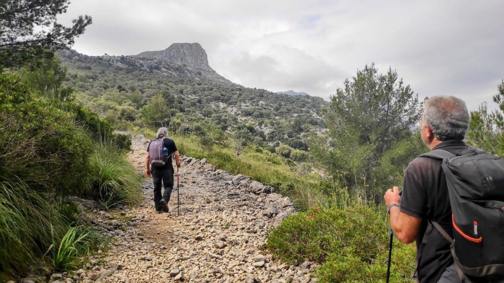 Dos senderistas en un camino de montaña con paisaje verde y rocoso al fondo.