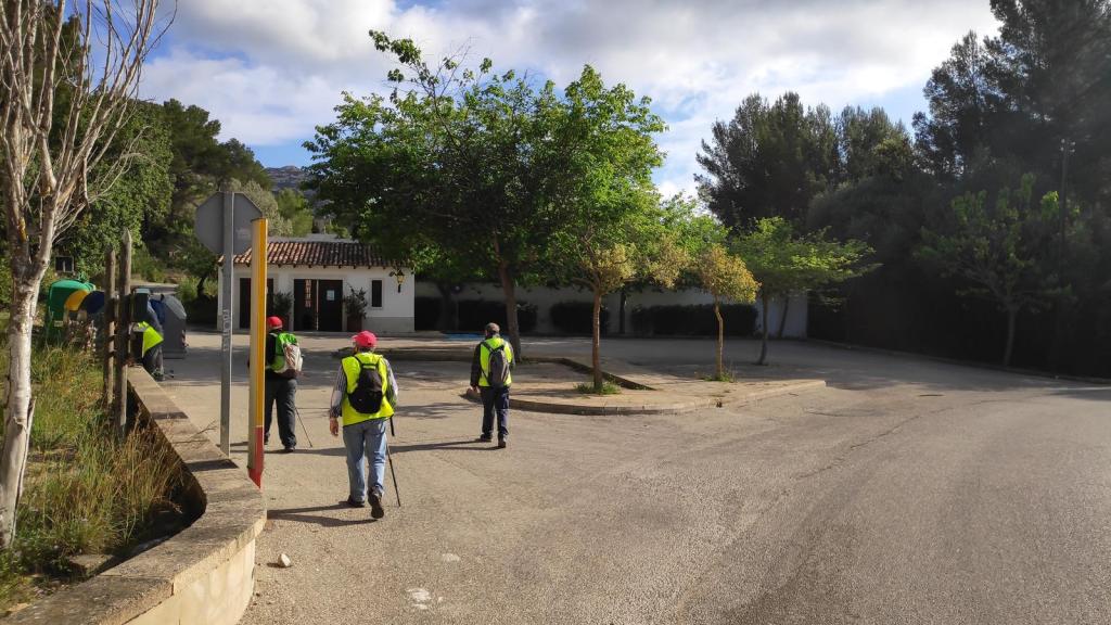 Un grupo de excursionistas con ropa de senderismo y chalecos reflectantes caminan hacia un cruce de caminos rodeado de vegetación en un entorno montañoso.