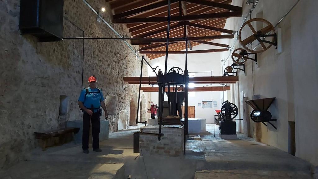 Interior de la Tafona de las Casas de Galatzó con maquinaria antigua, incluyendo molinos y herramientas agrícolas, mientras un visitante observa.