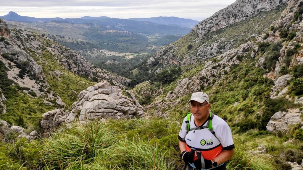 Hombre de pie en una senda montañosa rodeada de vegetación y rocas, con un paisaje montañoso al fondo.