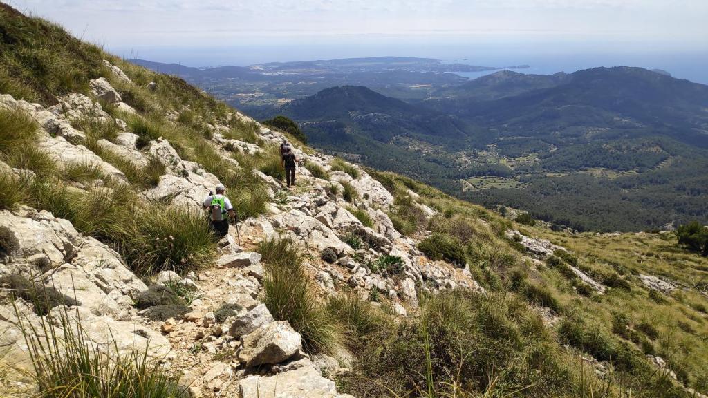 Vista de senderistas ascendiendo por un terreno rocoso y montañoso con vegetación, con el mar en el horizonte.