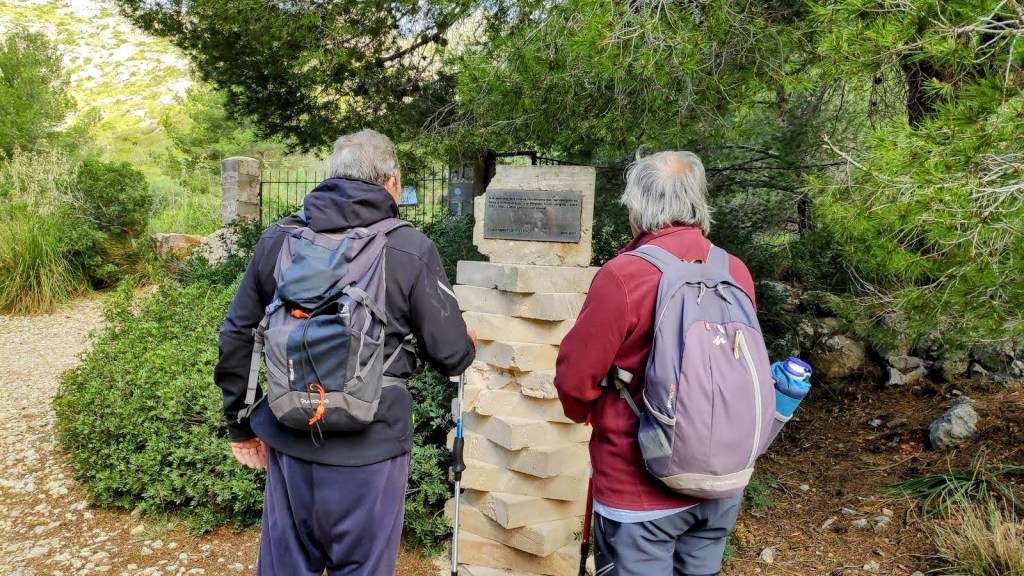 Dos personas observan un monolito con una placa conmemorativa en un camino rodeado de naturaleza verde, cerca de un bosque de pinos.