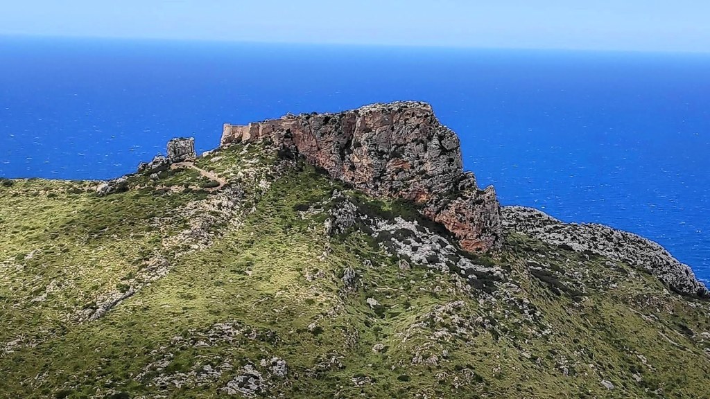 Vista panorámica del Castell del Rei con acantilados y mar de fondo, mostrando una formación rocosa prominente cubierta de vegetación.