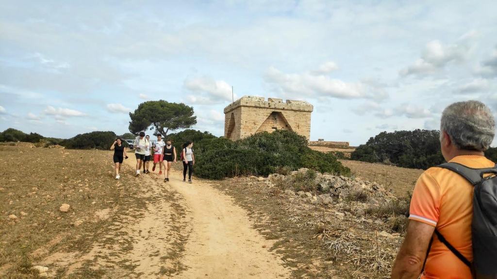 Grupo de personas caminando por un sendero hacia el Castell de Sa Punta de n'Amer, en un entorno natural con vegetación y cielo nublado.