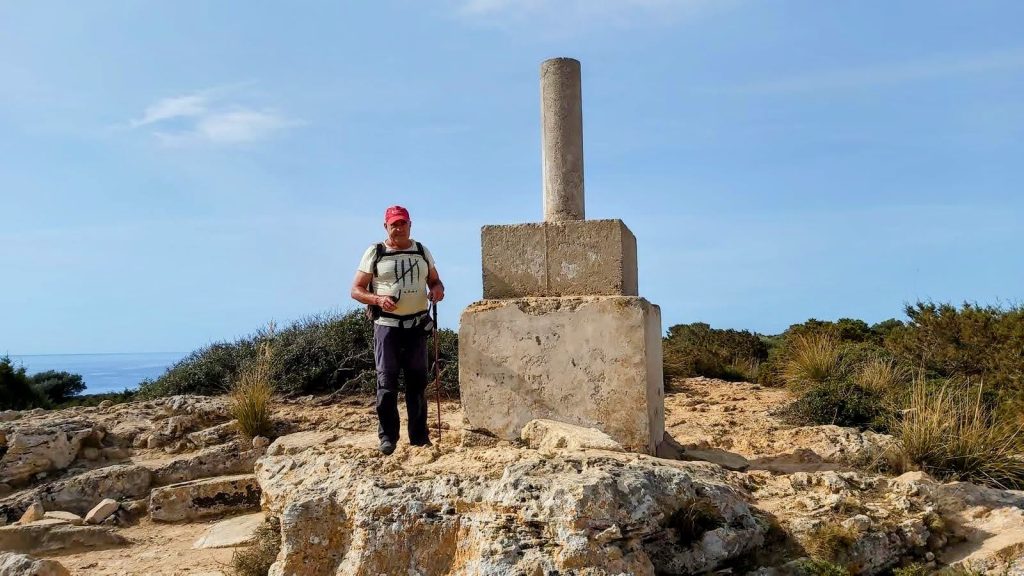 Hombre de pie junto a un vértice geodésico en Punta de n'Amer, con el mar y la vegetación al fondo.