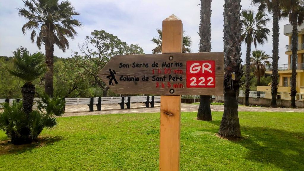 Cartel señalizador en un área verde que indica direcciones hacia 'Son Serra de Marina' y 'Colònia de Sant Pere', con un fondo de palmeras y un edificio al lado.
