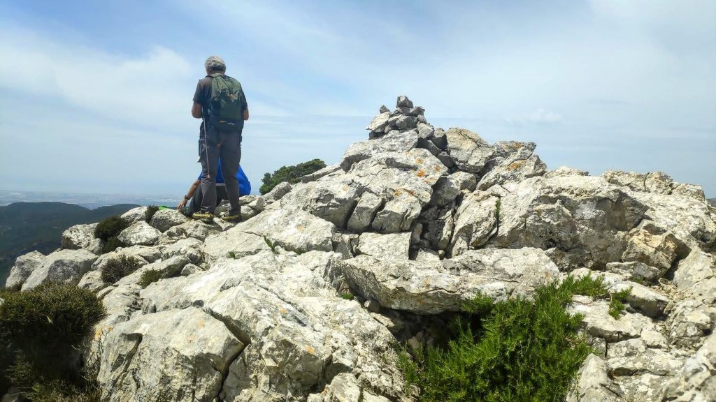 Persona de espaldas en la cima del Puig des Coll des Jou, rodeada de rocas y vistas al paisaje montañoso.