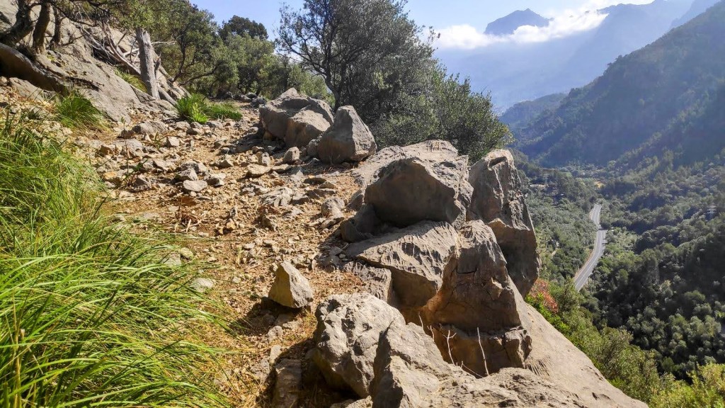 Sendero rocoso en una montaña rodeado de vegetación, con vistas panorámicas del valle y una carretera en el fondo.