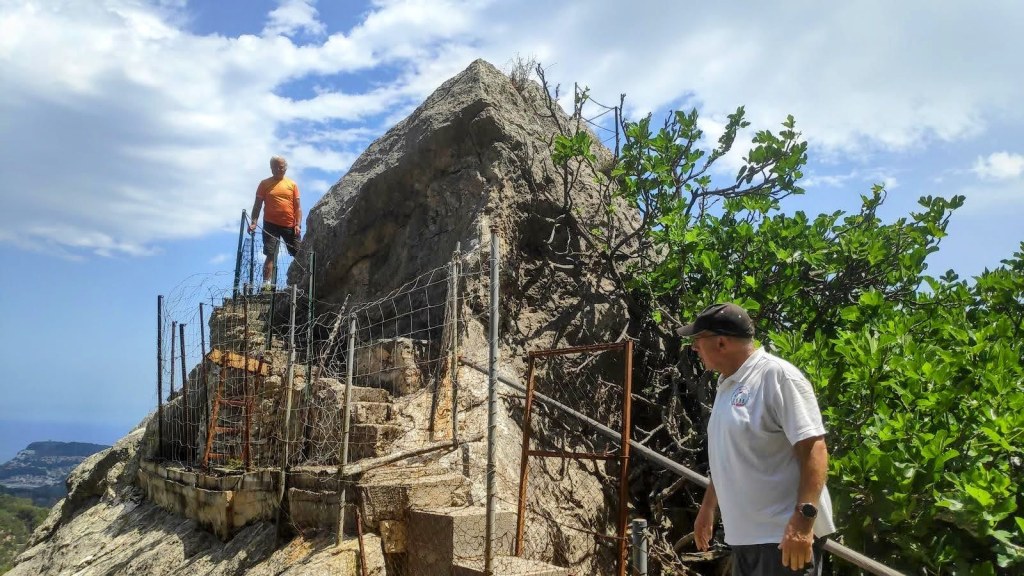 Dos personas en un camino de montaña, una está en una roca alta con una barandilla de protección y la otra se encuentra en el nivel inferior. Al fondo, se puede ver el paisaje de montañas y el mar.