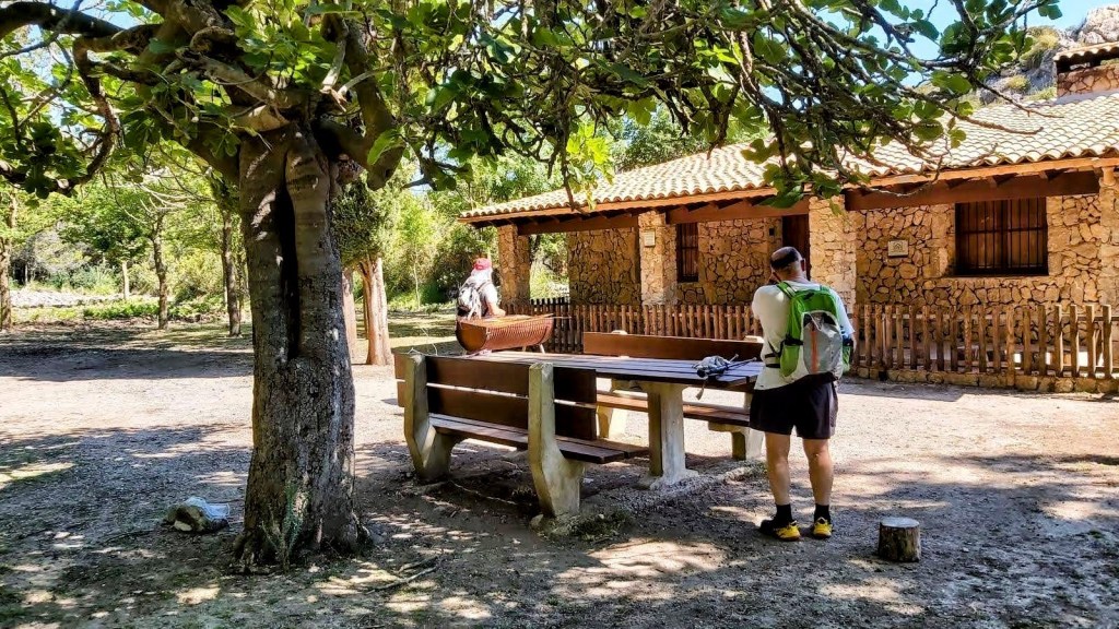 Un par de personas en el Refugio de Lavanor, con una mesa de picnic de madera y una cabaña de piedra al fondo, rodeados de árboles.