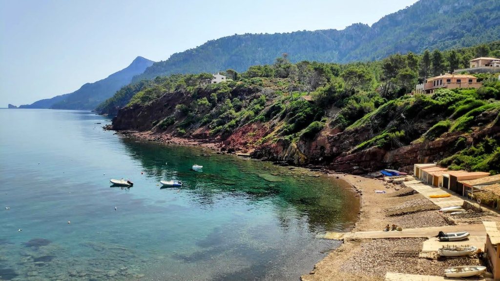 Vista panorámica del puerto des Canonge, con aguas cristalinas y pequeñas embarcaciones en la orilla, rodeado de montañas y vegetación.
