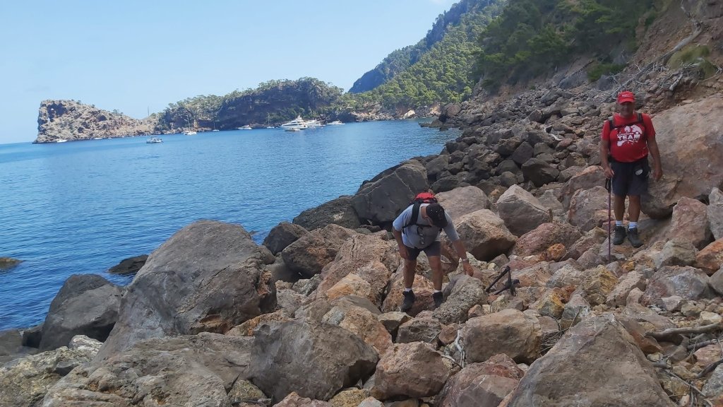 Dos excursionistas caminan entre rocas cerca de la costa en Caló de S'Estaca, con un mar azul al fondo y montañas verdes en el paisaje.