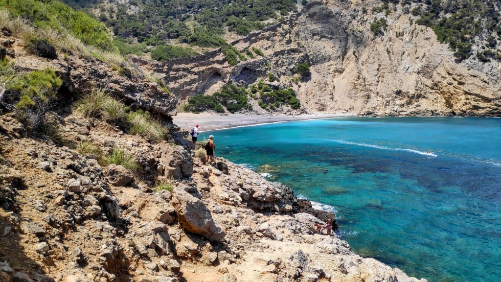 Cova Tancada-Playa del Coll Baix-Caminando por Mallorca Vista de la Playa del Coll Baix desde un acantilado, con varias personas en la orilla y aguas turquesas.