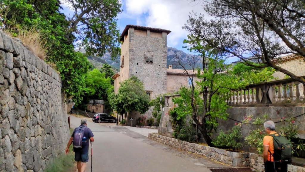 Un camino donde dos senderistas se dirigen hacia una torre de piedra de Son Marroig rodeada de vegetación y montañas, con un auto estacionado en la parte izquierda y muros de piedra a los lados.