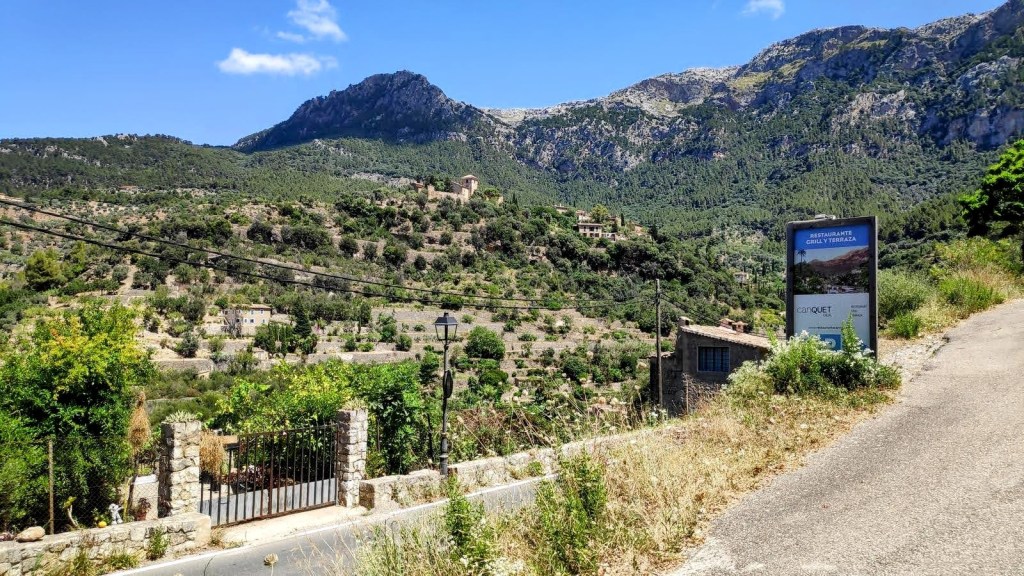 Vista de un paisaje montañoso en Deià, Mallorca, con vegetación abundante, casas distribuidas en la ladera y un cartel que indica la dirección hacia el restaurante Can Quet.