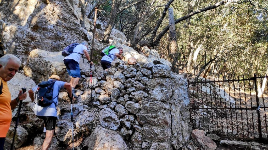 Grupo de senderistas ascendiendo por un camino rocoso con una muralla de piedra y un portón de hierro al fondo, rodeados de árboles en un bosque.