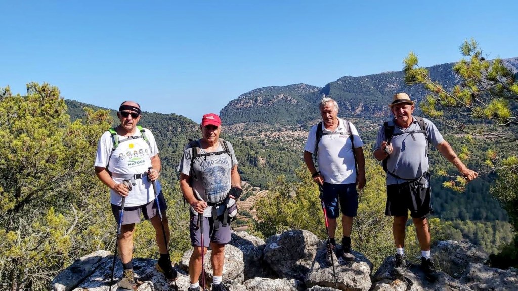 Cuatro hombres sonríen mientras posan en el Morro de Sa Bombarda, rodeados de un paisaje montañoso cubierto de vegetación, en un día soleado.