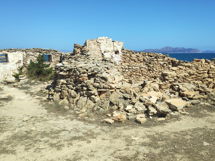 Ruinas de una antigua construcción de piedra junto al mar, con un cielo despejado y montañas al fondo.