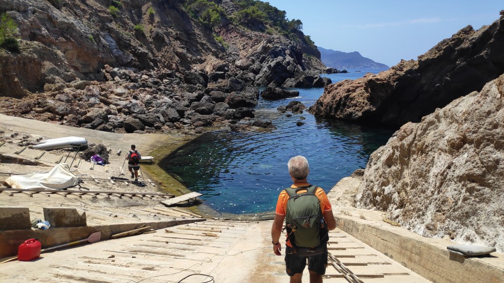 Hombre con mochila caminando hacia el  Caló o Port de S’Estaca rodeada de rocas en Mallorca, con mar en calma y cielos despejados.