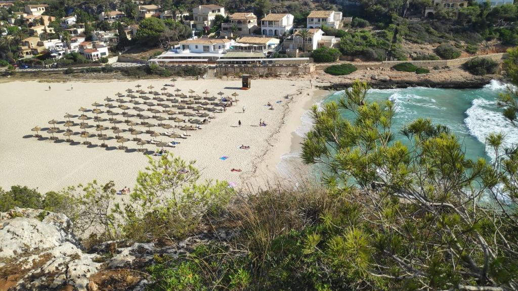 Vista aérea de Cala Romántica, con una playa de arena blanca, sombrillas dispuestas en filas y casas cercanas en una colina.