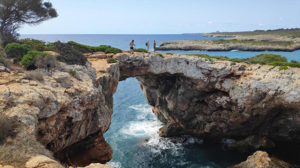 Vista de un puente natural formado por rocas sobre el mar, con personas de pie en la cima y un paisaje costero al fondo.