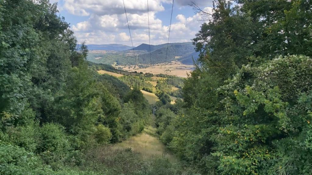 Vista panorámica del paisaje montañoso con árboles frondosos y un valle en el fondo, mostrando un cielo nublado y líneas eléctricas en el horizonte.