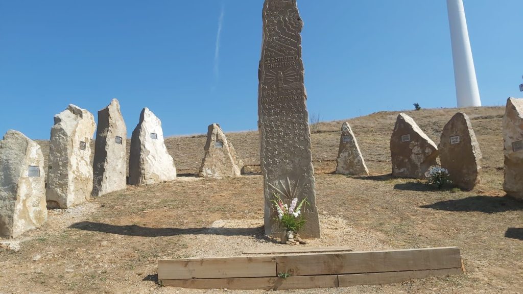 Un monumento con inscripciones y varios monolitos alrededor, ubicado en un paisaje árido bajo un cielo claro y soleado.