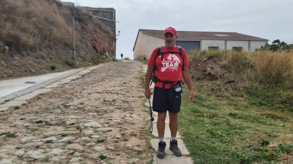 Camino de Santiago-Etapa 5: PuentelaReina-Estella - Caminando por Mallorca Hombre caminando por la calzada romana en la ruta del Camino de Santiago, llevando mochila y bastón, con un edificio al fondo.