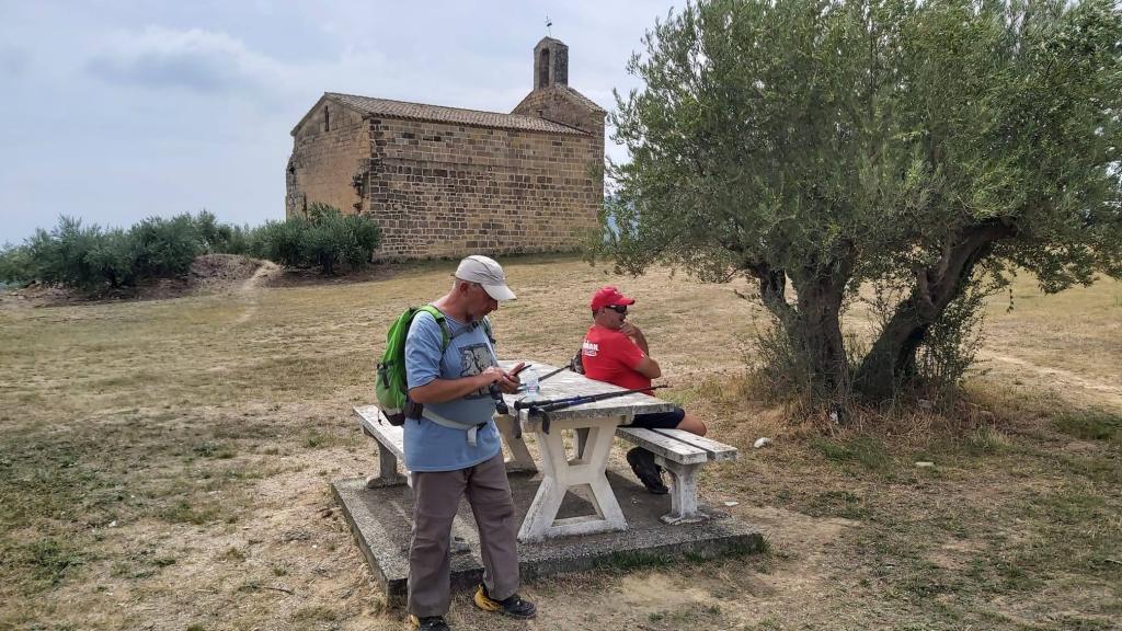 Camino de Santiago-Etapa 5: PuentelaReina-Estella - Caminando por Mallorca Dos hombres sentados en un banco de picnic frente a la Ermita de San Miguel, rodeados de un paisaje seco con un olivo al lado.