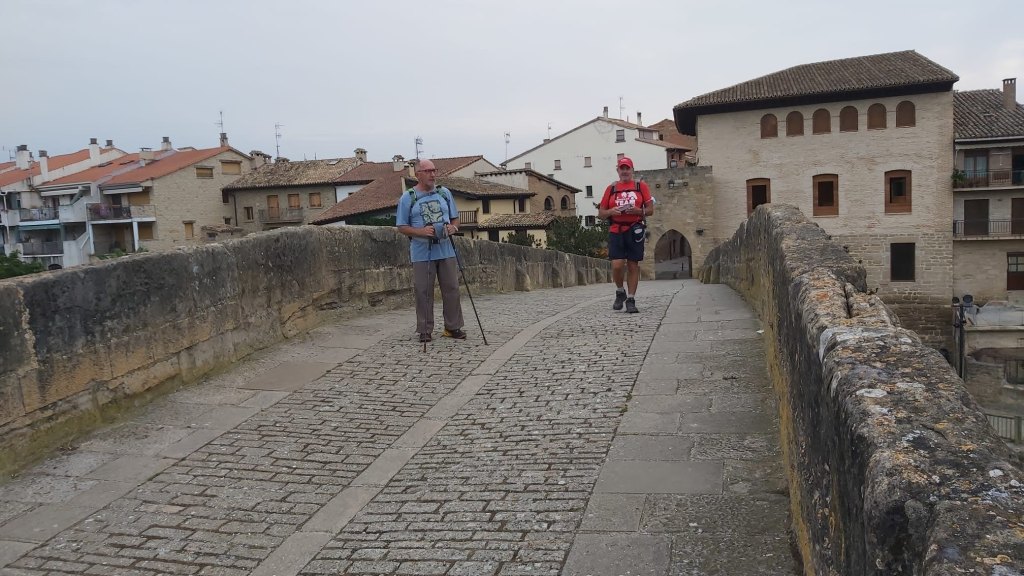 Dos caminantes en el Puente de los Peregrinoscon edificios históricos al fondo, durante la etapa 5 del Camino de Santiago.