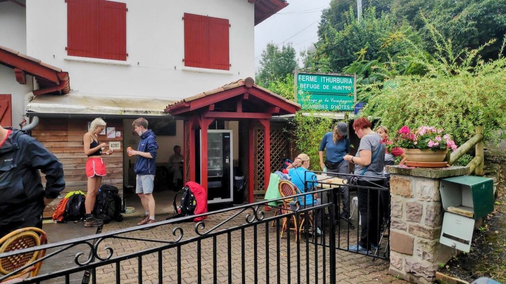 Grupo de personas en el refugio de Huntto, donde algunos están organizando sus pertenencias y otros están sentados en el exterior. Se puede ver la fachada del refugio con un cartel que indica su nombre.