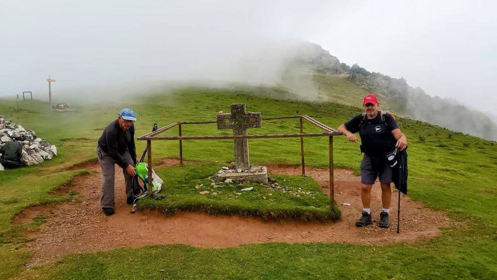 Dos peregrinos posando junto a la cruz de thibault en un entorno montañoso, con niebla y paisaje verde alrededor.