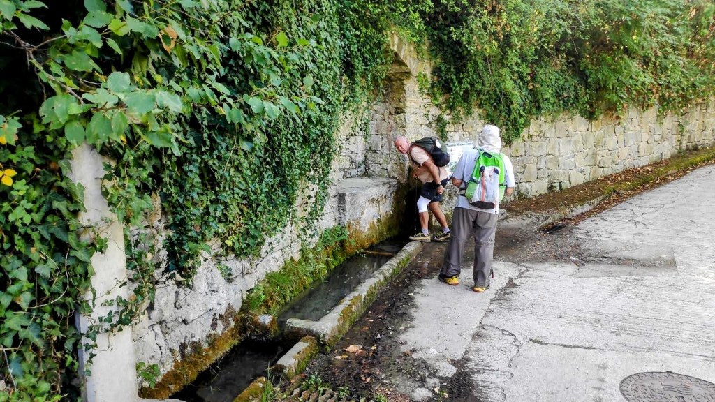 Dos personas llenando agua en la Fuente de Irotz. rodeada de vegetación y una pared de piedra, durante la ruta Zubiri-Pamplona del Camino de Santiago.