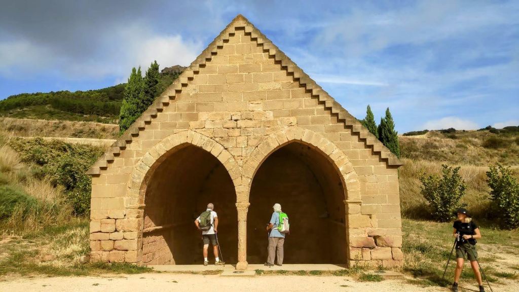 Vista de la Fuente de los Moros, de estilo medieval con arcos en su entrada, donde dos personas están de pie frente a él, en un entorno rural con vegetación y colinas al fondo.