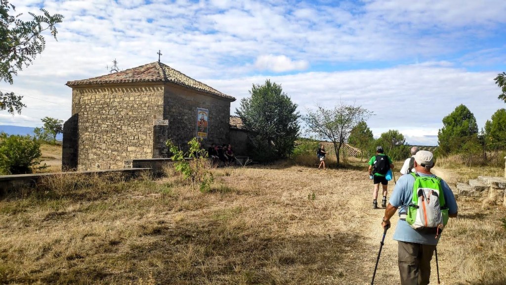 Grupo de caminantes en el Camino de Santiago, acercándose a la Ermita de la Virgen del Poyoen un entorno rural, con árboles y vegetación alrededor y un cielo parcialmente nublado.