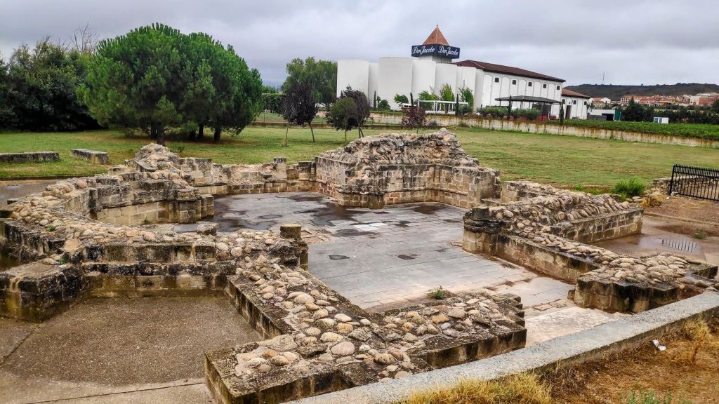Ruinas de un antiguo edificio con piedras dispuestas en forma circular, rodeadas de césped y árboles, en un paisaje rural con un edificio al fondo.