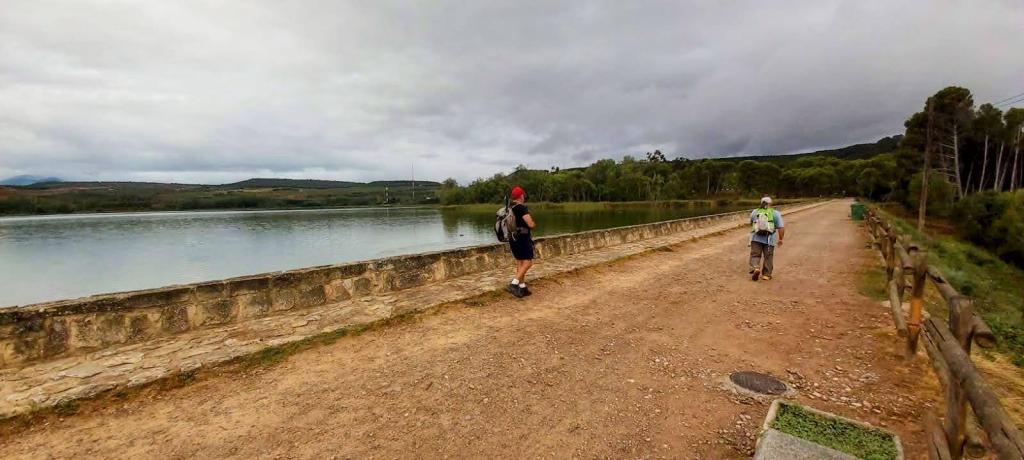 Dos personas caminando por un sendero junto a un embalse, con vegetación al lado y un cielo nublado.