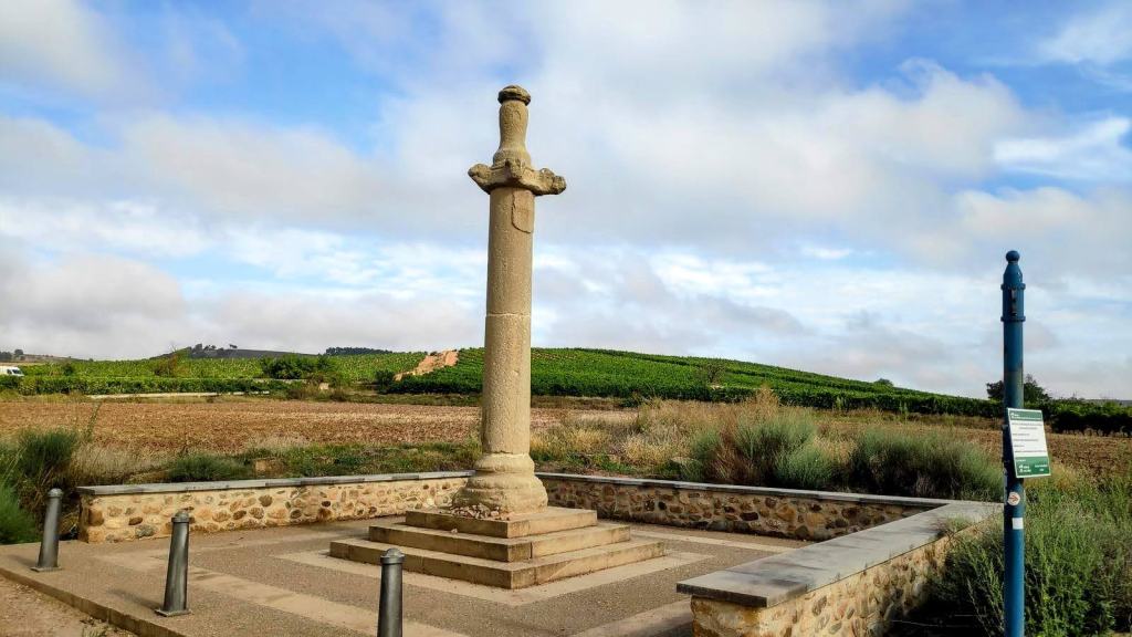 Columna de piedra con un crucero en la cima, ubicada en un paisaje agrícola con viñedos al fondo y un cielo parcialmente nublado.