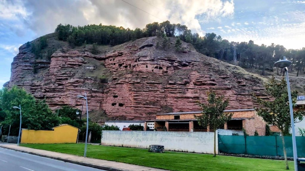 Vista de las Cuevas  de Nájera en un acantilado de roca roja con árboles en la parte superior, junto a una vía pavimentada y edificios en la base.