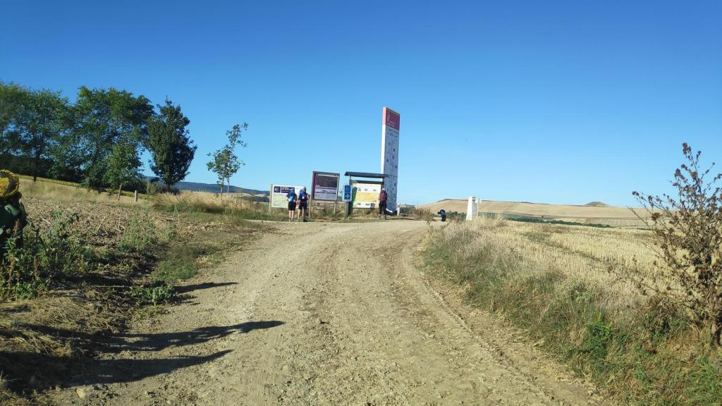 Frontera La Rioja-Castilla León, mostrando un camino de tierra y un panel informativo con multitudes de árboles y campos de cereal al fondo.