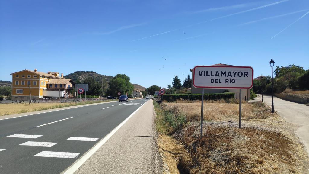 Cartel de bienvenida al pueblo de Villamayor del Río, con una carretera y una vivienda amarilla al fondo, bajo un cielo despejado.